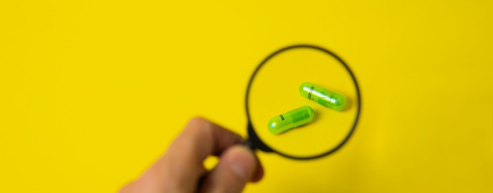 From above of crop unrecognizable male holding magnifying glass over green pills on yellow background in studio — The Ultimat