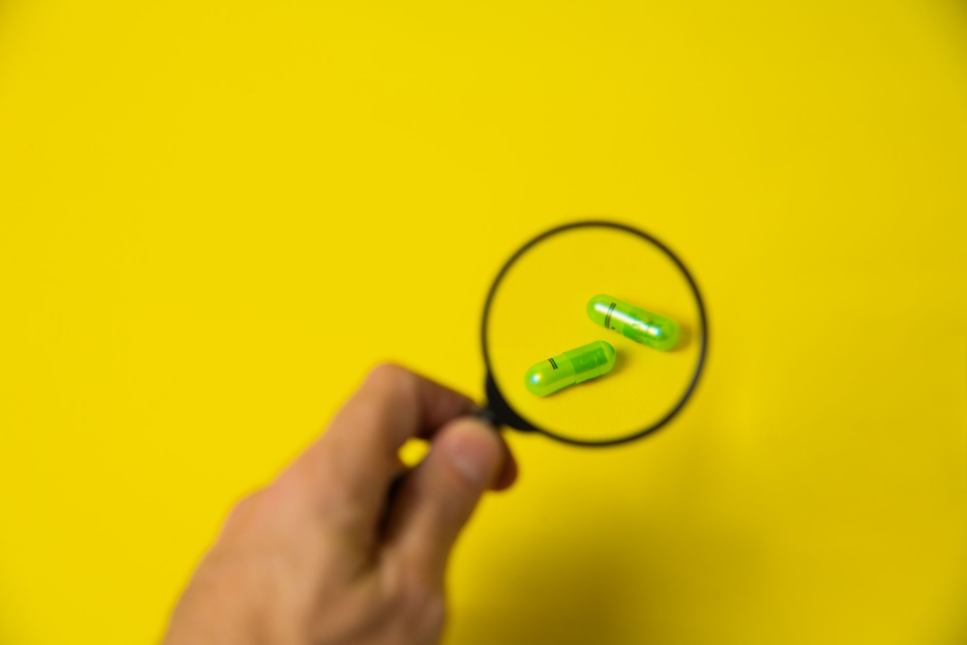 From above of crop unrecognizable male holding magnifying glass over green pills on yellow background in studio — The Ultimat